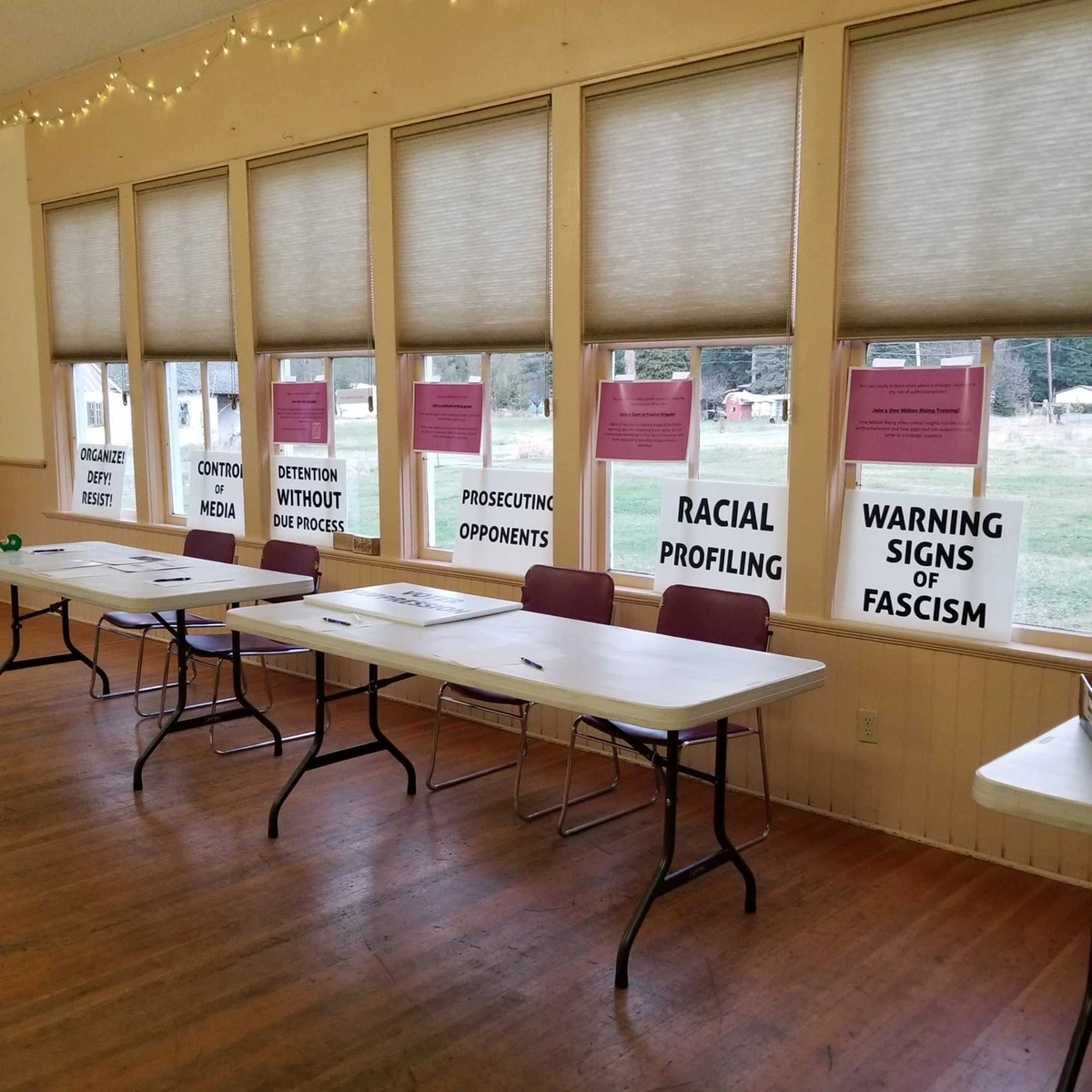 A row of sign up stations with pink sheets advertising activism opportunities and white signs reading "Organize! Defy! Resist!", "Control of Media", "Prosecuting Opponents", "Racial Profiling", and "Warning Signs of Fascism"